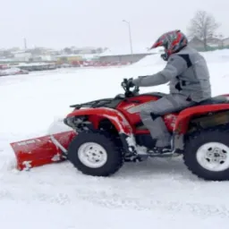 Czerwony quad Yamaha z pługiem śnieżnym odśnieża teren pokryty grubą warstwą śniegu, kierowca w kasku i szarym kombinezonie.