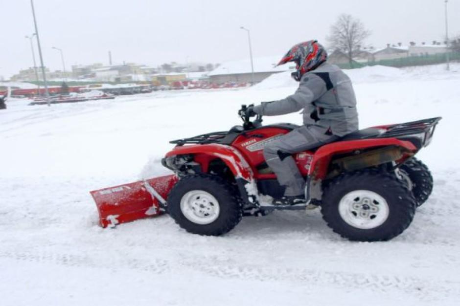 Czerwony quad Yamaha z pługiem śnieżnym odśnieża teren pokryty grubą warstwą śniegu, kierowca w kasku i szarym kombinezonie.