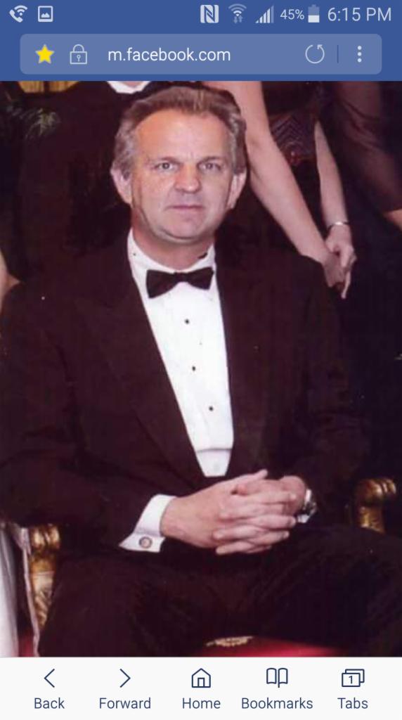 Mężczyzna w czarnym smokingu z muszką, siedzący na zdobionym krześle, z dłońmi złożonymi na kolanach, otoczony przez częściowo widoczne osoby w tle.