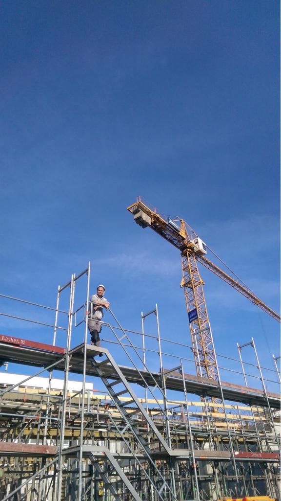 Pracownik w kasku i kombinezonie opiera się o barierkę rusztowania na tle błękitnego nieba i żurawia budowlanego, widok z dołu.