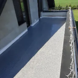 Wykończony dach z lukarnami i balustradą, pokryty jasnoszarą, chropowatą powłoką z widocznymi detalami wykończeniowymi i fragmentem krajobrazu w tle.