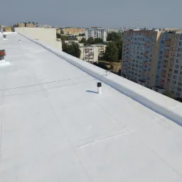 Biały dach budynku wielorodzinnego po renowacji, widok z góry na tle zabudowy miejskiej w słoneczny dzień. Nowa, jasna powłoka hydroizolacyjna.