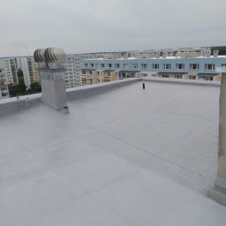 ARS-DACHY - Szary, płaski dach budynku wielorodzinnego z dwoma turbowentylatorami. W tle widoczne inne budynki i pochmurne niebo.