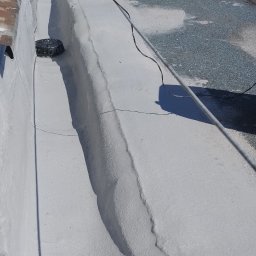 ARS-DACHY - Biała, płaska powierzchnia dachu po renowacji z widocznym wpustem kanalizacyjnym i rolkami papy w tle. Prace dekarskie w słoneczny dzień.