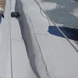 Biała, płaska powierzchnia dachu po renowacji z widocznym wpustem kanalizacyjnym i rolkami papy w tle. Prace dekarskie w słoneczny dzień.