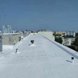 Biały dach pokryty powłoką hydroizolacyjną na tle miejskiego krajobrazu z budynkami i niebem. Widoczne elementy wentylacyjne i kominowe.