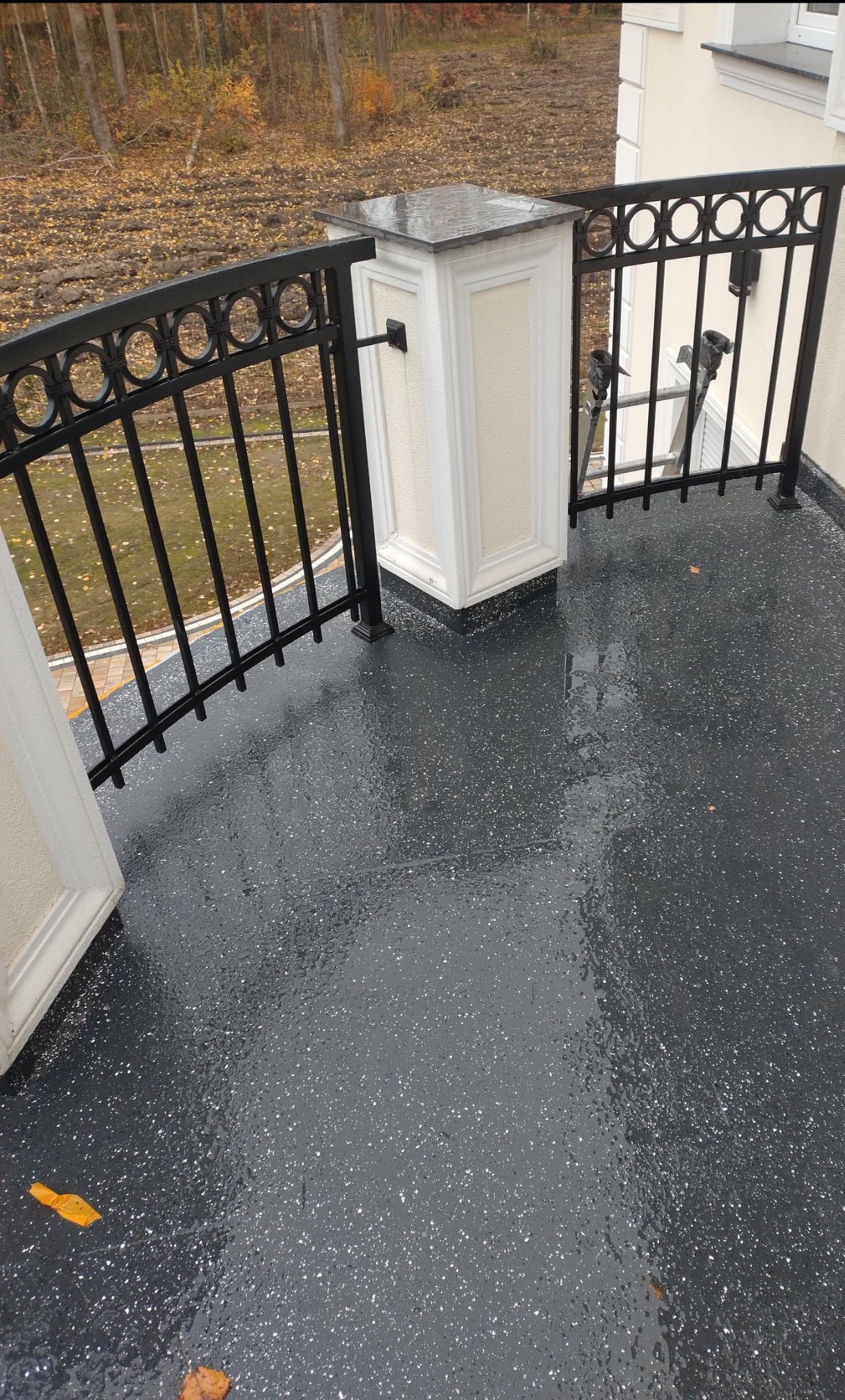 Balkon z czarną, błyszczącą posadzką z białymi drobinkami, otoczony czarną balustradą z dekoracyjnymi kółkami i kremowymi filarami. Widoczne jesienne liście na posadzce i drzewa w tle.