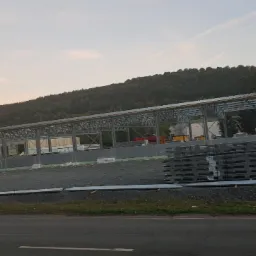 Montaż konstrukcji stalowej hali z widocznymi elementami płyt warstwowych i stosem blach, na tle zalesionego wzgórza i zachodzącego słońca.