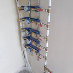Pionowo zamontowany zestaw wodomierzy z zaworami odcinającymi i rurami z tworzywa na metalowej szynie montażowej, widok z dołu.