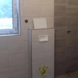 Świeżo wykafelkowana łazienka z zabudowaną toaletą i żółtymi przyłączami hydraulicznymi, widok na okno z białymi ramami.