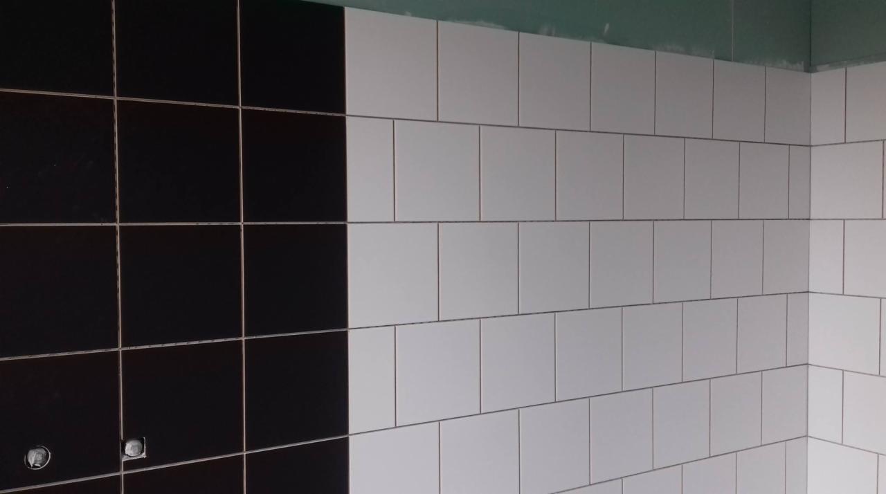 Ściana wyłożona białymi i czarnymi, kwadratowymi płytkami ceramicznymi, widoczne puszki elektryczne, szara ściana nad płytkami.