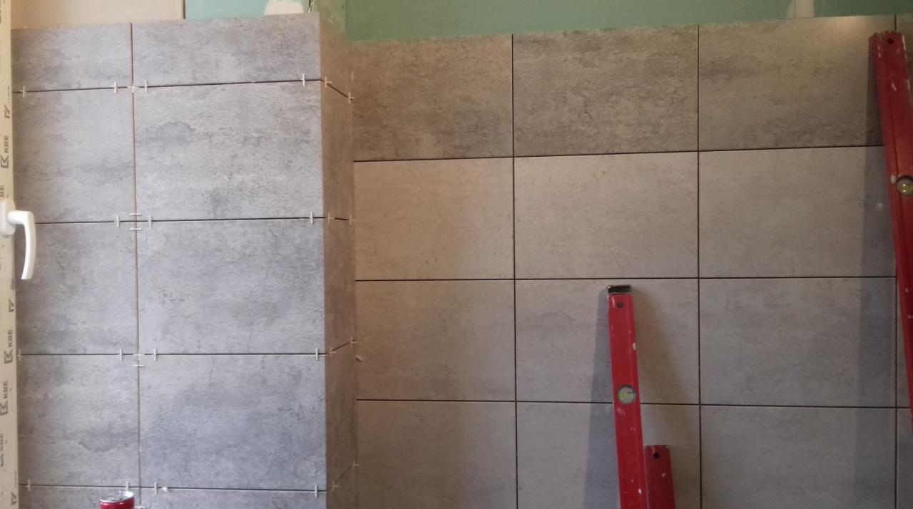 Ściana w trakcie układania płytek ceramicznych w łazience, widoczne krzyżyki dystansowe, poziomica i fragment ościeżnicy.