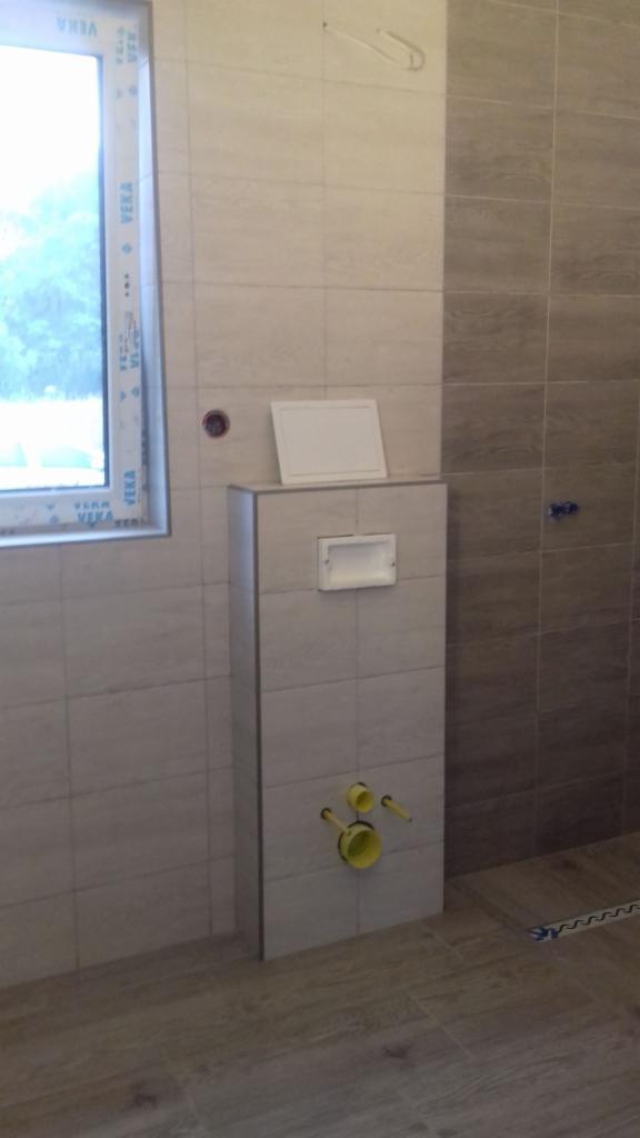 Świeżo wykafelkowana łazienka z zabudowaną toaletą i żółtymi przyłączami hydraulicznymi, widok na okno z białymi ramami.