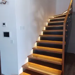 Nowoczesne schody drewniane z podświetleniem LED stopni i metalową balustradą o ażurowym wzorze, zamontowane w jasnym wnętrzu z widocznymi panelami sterowania na ścianie.