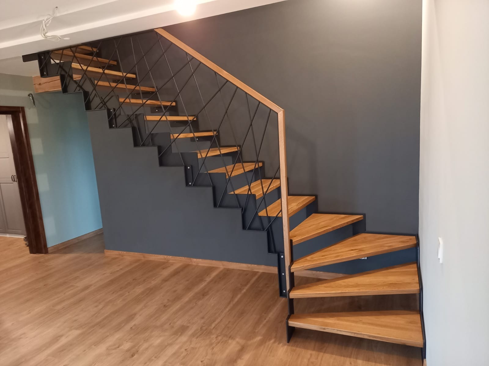Nowoczesne schody z drewnianymi stopniami i czarną, geometryczną balustradą z metalu na tle szarej ściany.
