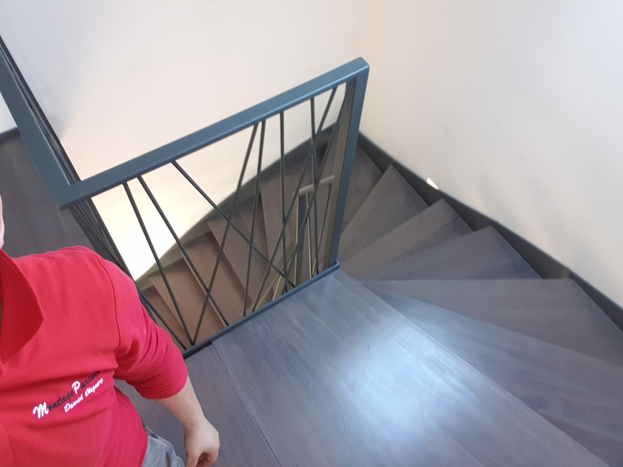 Widok z góry na drewniane schody z metalową balustradą o minimalistycznym designie, z widoczną osobą w czerwonej bluzie po lewej stronie kadru.