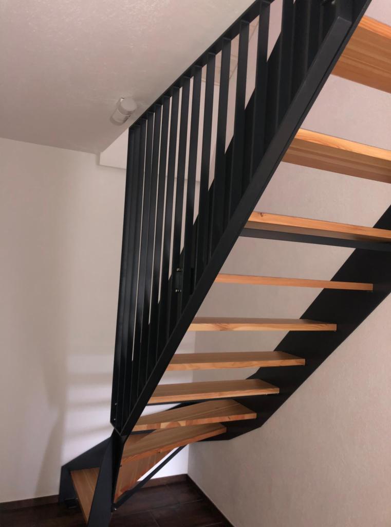 Nowoczesne schody metalowe z drewnianymi stopniami, czarna balustrada z pionowych prętów, widok od dołu, oświetlenie sufitowe w tle.