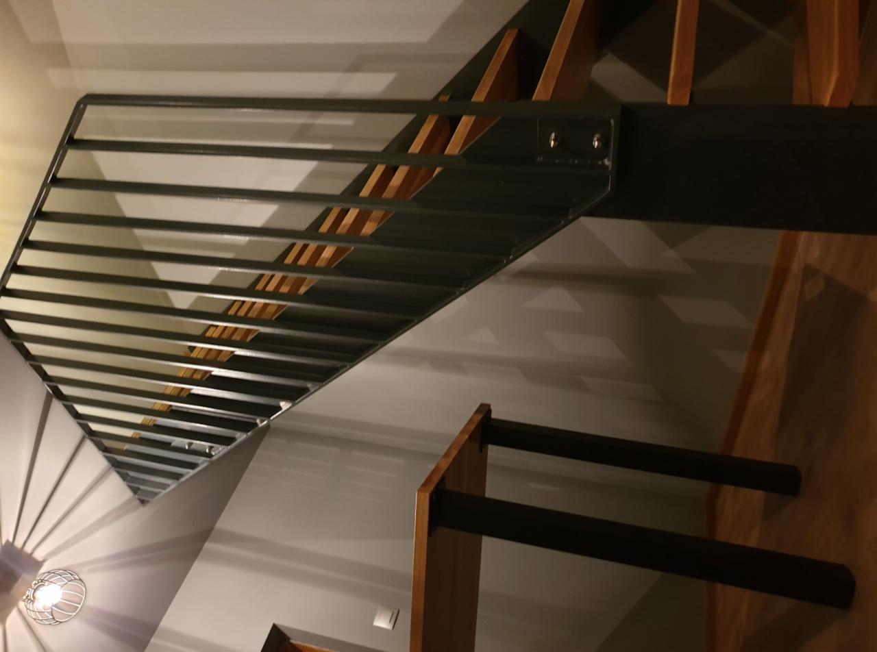 Schody metalowe z drewnianymi stopniami i metalową balustradą, widok z dołu, oświetlone ciepłym światłem z lampy, cień rzucany na ścianę.