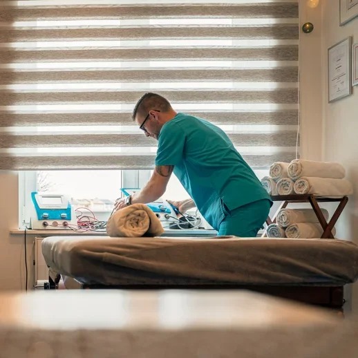 Mężczyzna w zielonym uniformie, pochylony nad pacjentem leżącym na stole do masażu, podłączony do aparatury do elektroterapii, w tle okno z żaluzjami i półka z ręcznikami.