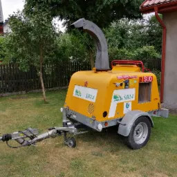 Żółta, mobilna rębarka do gałęzi marki Saelen SB14 z widocznym dyszlem i kołami, stojąca na trawniku przed budynkiem.