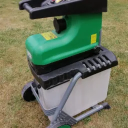 Elektryczny rozdrabniacz do gałęzi w kolorze zielono-czarnym, z przezroczystym pojemnikiem na odpady, stojący na trawie.