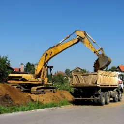 Żółta koparka nabiera ziemię i wysypuje ją do skrzyni ciężarówki na tle zabudowy jednorodzinnej w słoneczny dzień.