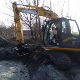 Żółta koparka JCB z operatorem w kabinie, pracująca w błotnistym terenie z fragmentami gałęzi i korzeni, widoczne logo Barton Manor Developments Ltd na ramieniu maszyny.