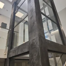 Szklana winda wewnątrz budynku z betonowymi elementami konstrukcyjnymi i oświetleniem sufitowym, widok z dołu.