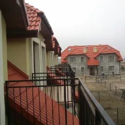 Widok z balkonu na budowę osiedla domów jednorodzinnych z czerwonymi dachami, w tle pracownicy budowlani i ogrodzenie posesji.