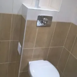 Nowoczesna toaleta podwieszana z chromowanym przyciskiem spłukiwania, beżowe płytki ścienne i podłogowe z fugą w kolorze ecru, biała półka nad toaletą z rolką po papierze toaletowym.