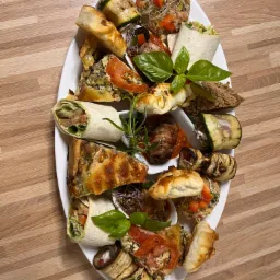 Elegancka biała taca z różnorodnymi przekąskami: tortille z warzywami, mini quiche, roladki z grillowanego bakłażana i cukinii, ciastka francuskie, kulki z kiełkami, udekorowane świeżymi ziołami.