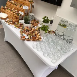 Stół cateringowy przykryty białym obrusem, z drewnianą kaskadą pełną kanapek i tartinek z różnymi dodatkami, szklanki ustawione w rzędach, dwa szklane dystrybutory wody z cytryną i miętą, doniczki...