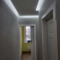 Szare wnętrze korytarza z podwieszanym sufitem z oświetleniem LED, widok na okno z kratą i drzwi z białymi ościeżnicami.