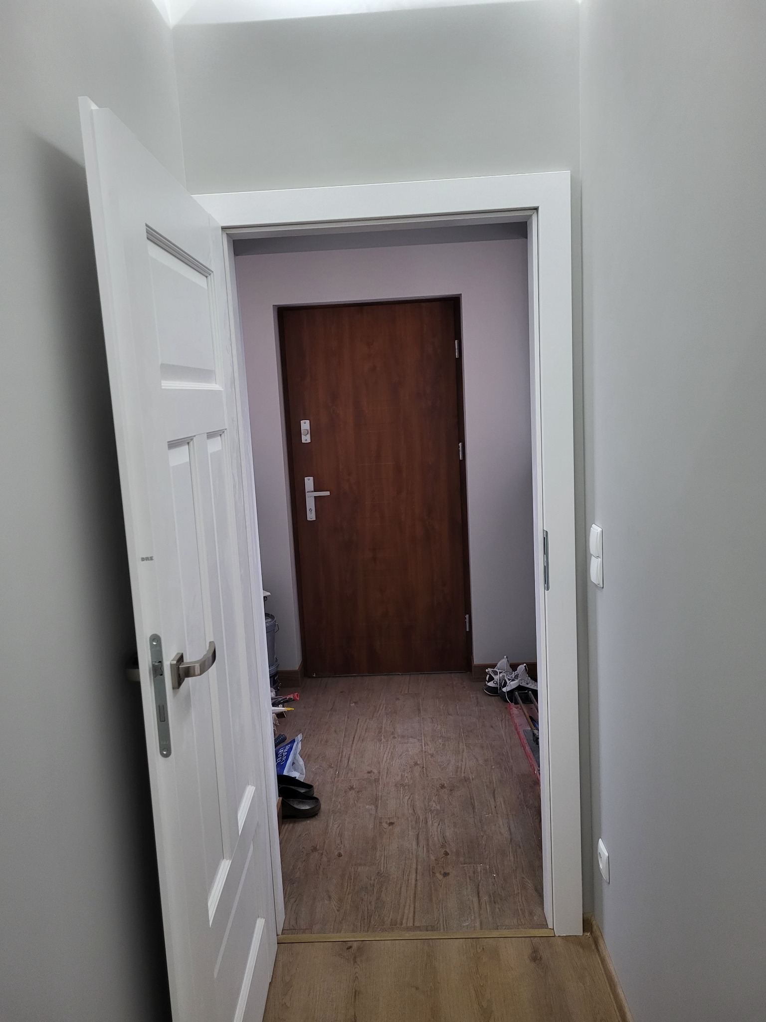 Widok na korytarz z otwartymi, białymi drzwiami wewnętrznymi, ukazujący brązowe drzwi wejściowe w głębi oraz fragment podłogi z laminatu i pozostawione buty.