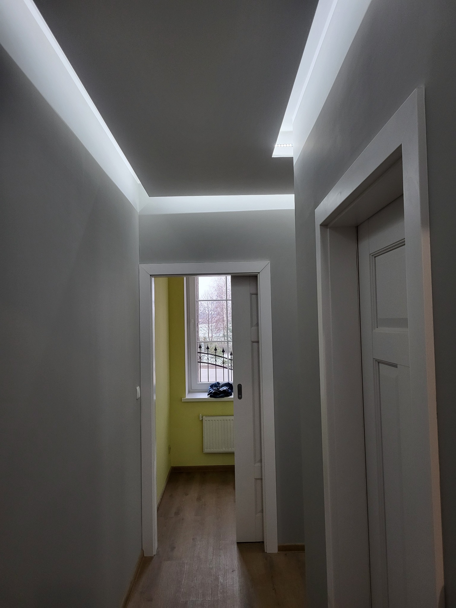 Szare wnętrze korytarza z podwieszanym sufitem z oświetleniem LED, widok na okno z kratą i drzwi z białymi ościeżnicami.