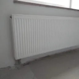 Biały, panelowy grzejnik zamontowany na ścianie w surowym stanie, widoczne ślady po instalacji hydraulicznej i niedokończone wykończenie pod oknem.