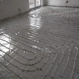 Białe rurki ogrzewania podłogowego ułożone spiralnie na folii z nadrukiem na przygotowanym podłożu w surowym pomieszczeniu z oknem.