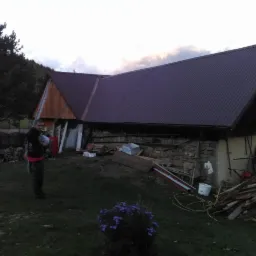 Dom z nowym, brązowym dachem z blachy trapezowej. Na ścianie sterta desek i drabina. Mężczyzna stoi na trawniku. Widok na góry i zachmurzone niebo.