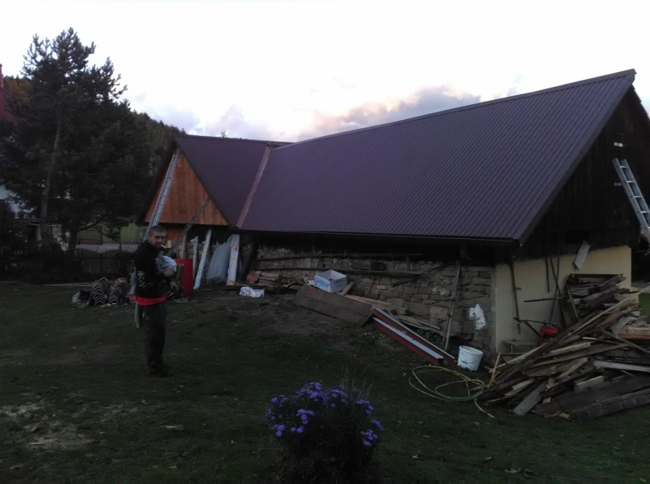 Dom z nowym, brązowym dachem z blachy trapezowej. Na ścianie sterta desek i drabina. Mężczyzna stoi na trawniku. Widok na góry i zachmurzone niebo.