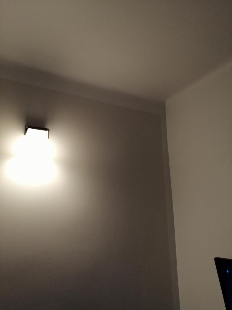 Ściana z zamontowanym, świecącym kinkietem o prostokątnej podstawie, oświetlającym biały sufit i fragment ściany w rogu pokoju.