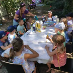 Dzieci siedzą przy stole w ogrodzie, uczestnicząc w zajęciach plastycznych pod okiem animatorki w fioletowej koszulce i rękawiczkach. Na stole znajdują się farby, pędzle, pojemniki i butelki z olejem.