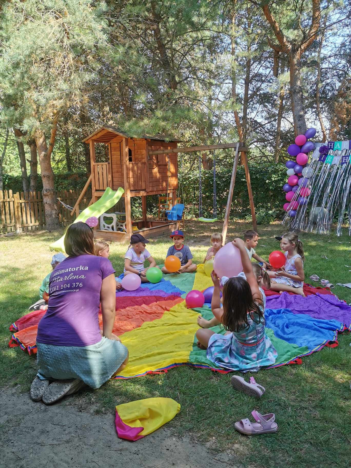 Grupa dzieci siedzi w kręgu na kolorowej chuście podczas zabawy z balonami na trawie, w tle drewniany domek na drzewie i dekoracje urodzinowe.