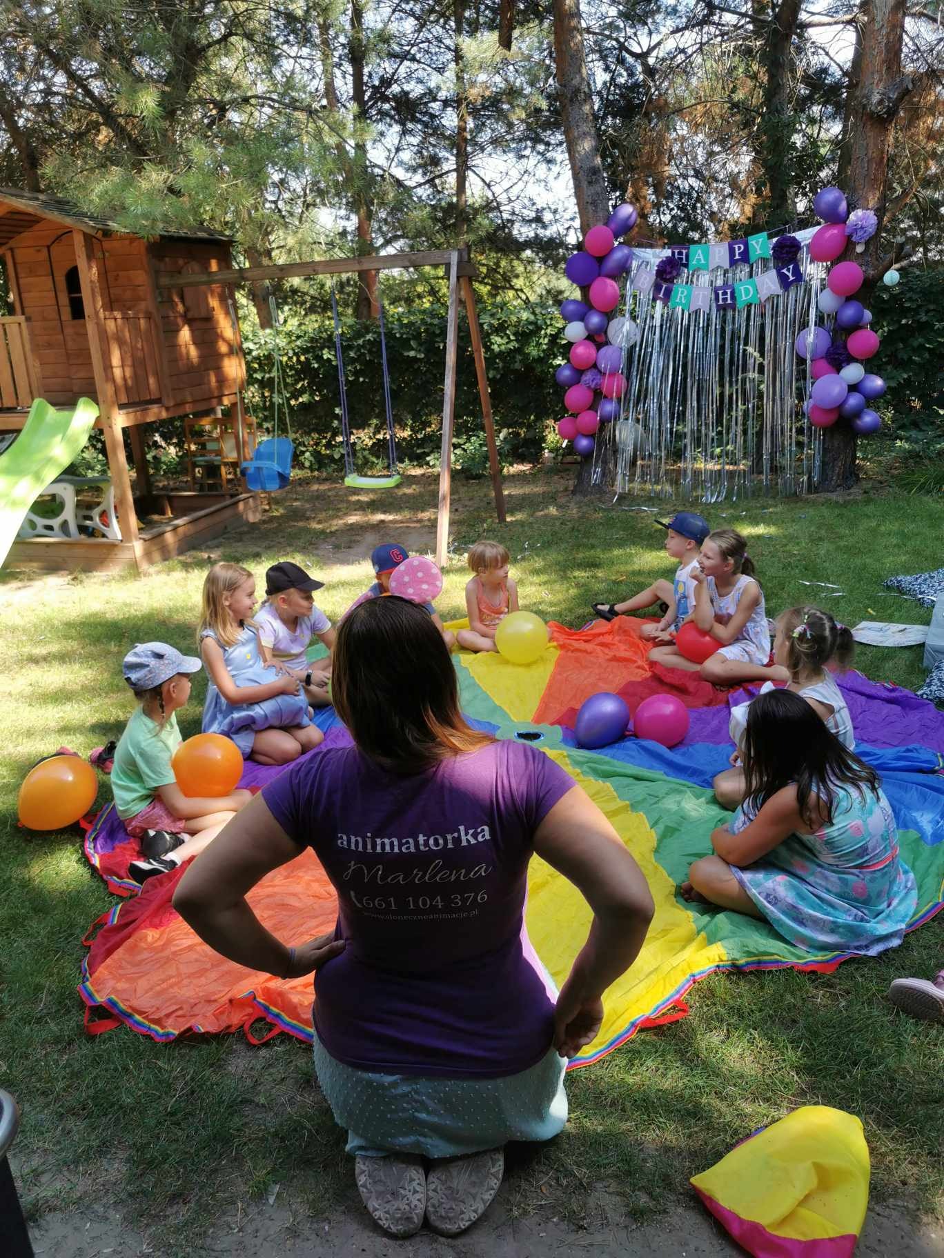 Grupa dzieci siedzi na kolorowej chuście animacyjnej na trawie podczas przyjęcia urodzinowego, w tle dekoracje z balonów i napis 'Happy Birthday', widoczna animatorka w fioletowej koszulce...