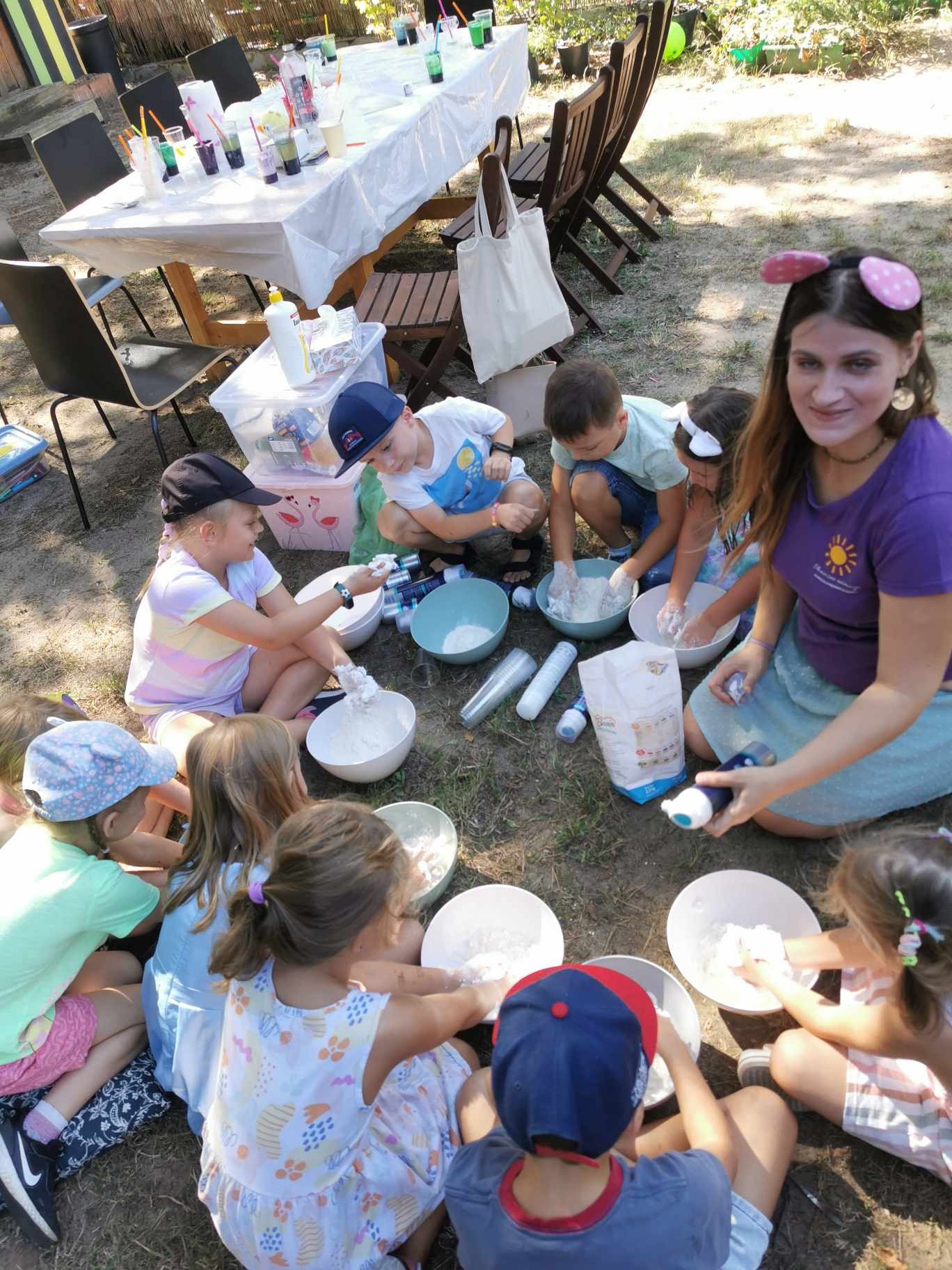 Grupa dzieci i animatorka siedząca na trawie, mieszają składniki w miskach, prawdopodobnie podczas warsztatów slime na świeżym powietrzu; na stole w tle napoje w kubkach.
