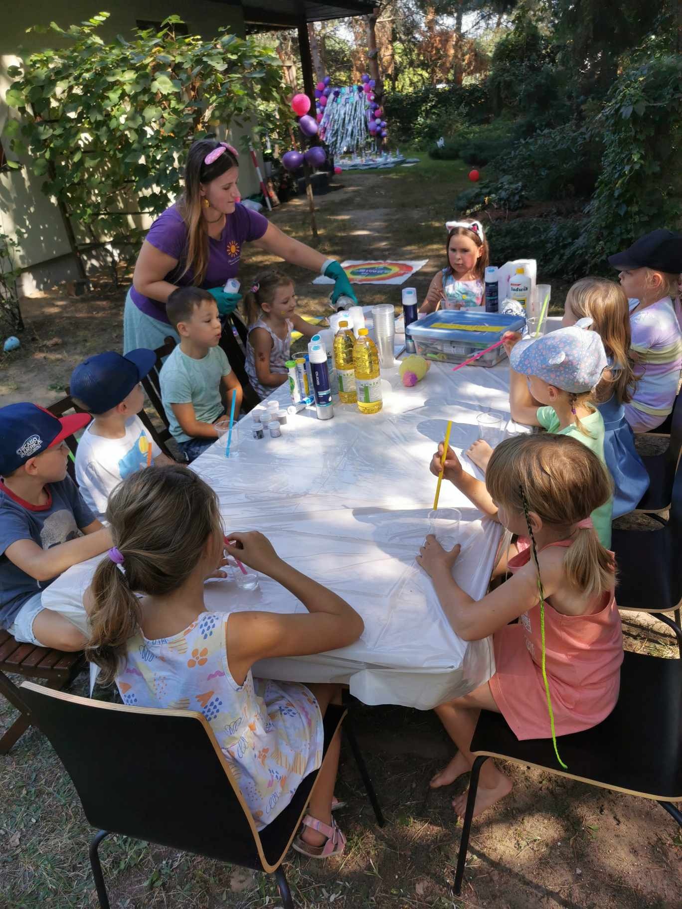 Dzieci siedzą przy stole w ogrodzie, uczestnicząc w zajęciach plastycznych pod okiem animatorki w fioletowej koszulce i rękawiczkach. Na stole znajdują się farby, pędzle, pojemniki i butelki z olejem.