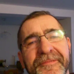 Portret mężczyzny w okularach, z lekkim uśmiechem, w pomieszczeniu z widocznymi meblami w tle.