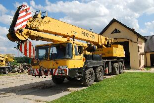 Żółty dźwig samochodowy z wysięgnikiem teleskopowym, zaparkowany na placu przed budynkiem gospodarczym. Widoczne detale konstrukcyjne i oznaczenia na dźwigu.
