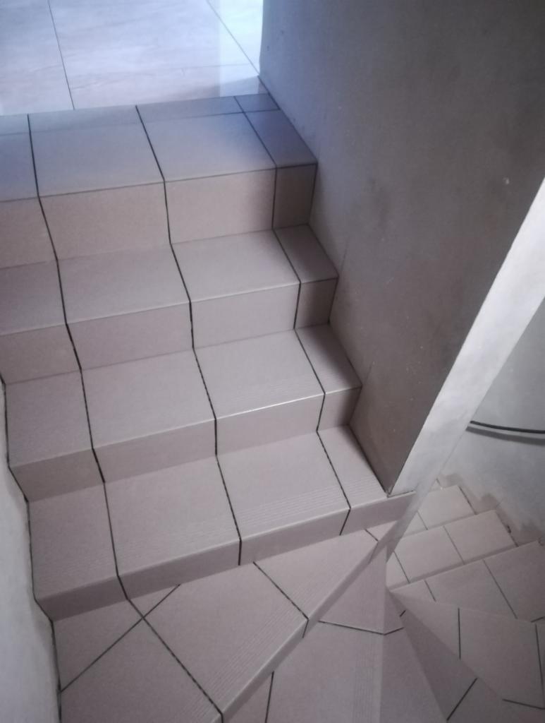 Wyłożone beżowymi płytkami schody z ciemnymi fugami, widok z góry, ściana w tle o surowym wykończeniu.