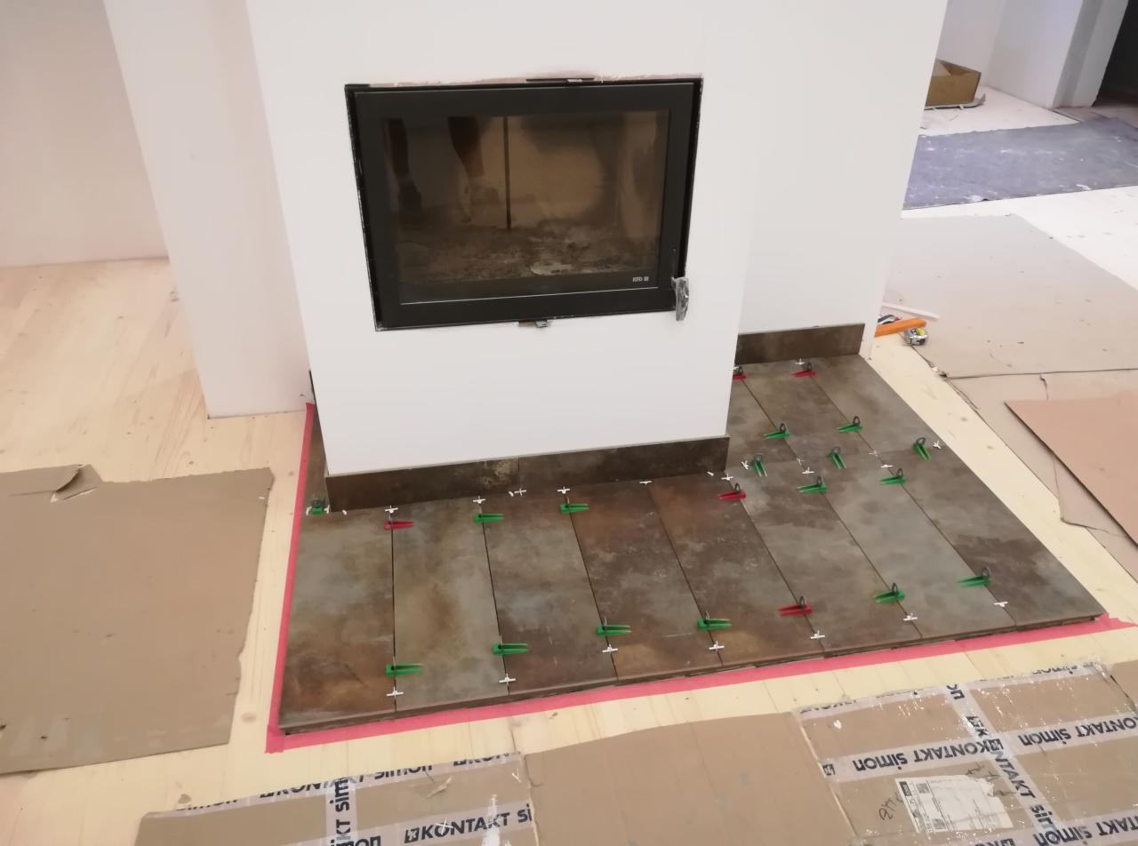 Montaż brązowych płytek pod kominkiem, widoczne zielone i czerwone kliny dystansowe, zabezpieczenie podłogi kartonem.