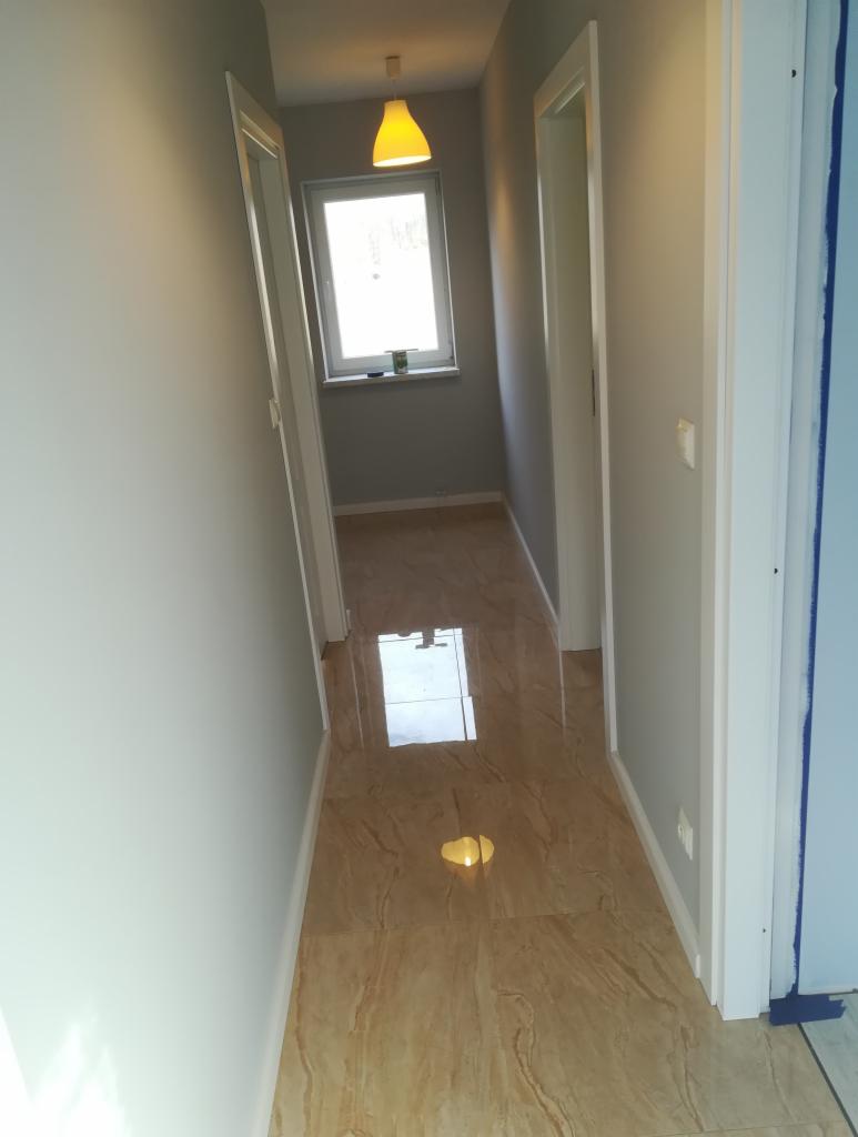 Korytarz po remoncie z beżowymi płytkami podłogowymi imitującymi marmur, szare ściany, białe ościeżnice i żółta lampa sufitowa. Na parapecie okna widoczny mały przedmiot.